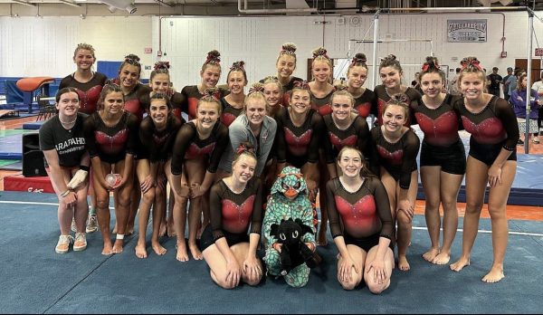 The team smiles after completing the first meet of the season. The team had their mascot, the dragon, with them to lift up their spirits. One of the gymnasts little brother dresses up in a costume to match the team. 